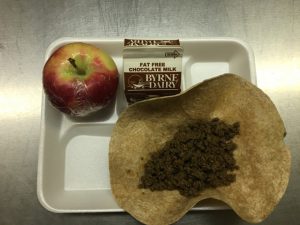 cafeteria tray with a taco shell, crumbled hamburger, chocolate milk and an apple