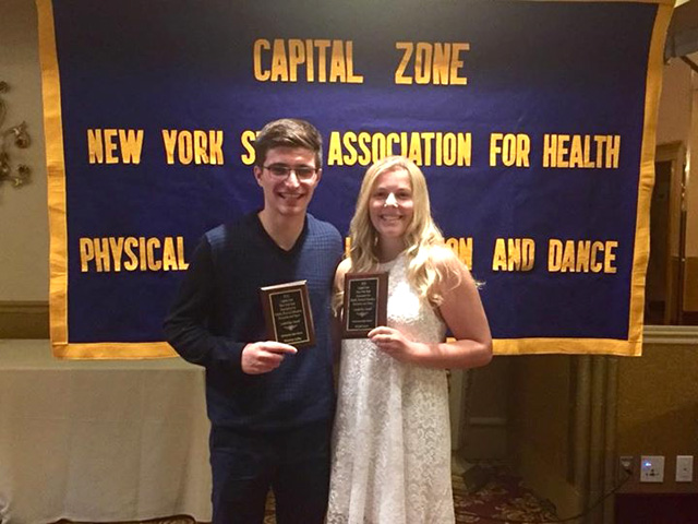 two winners hold small plaques while standing in front of Capital Zone banner