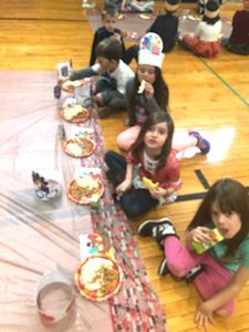 students sit crossed legged on gym floor eating from plates on tablecloths
