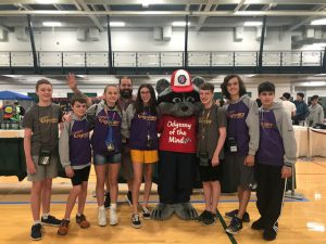 eight teens pose with the Odyssey of the Mind mascot, a raccoon dressed in a red t-shirt and red and white ball cap