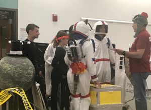 students in astronaut costumes talk with an adult wearing a red t-shirt