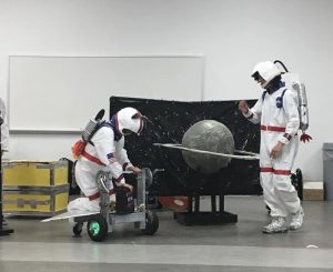 two students dressed as astronauts perform on stage with props that include a black backdrop and a planet with rings