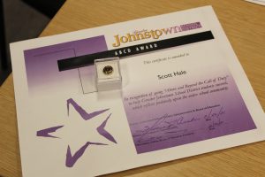 A certificate is shown in Johnstown colors, purple and gold, announcing Scott Hale as the recipient of an ABCD Award. An ABCD Award pin is shown on top of the certificate.