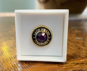 A commemorative pin is shown that has a purple stone in the middle, with the words "ABCD Award, Johnstown" on it.