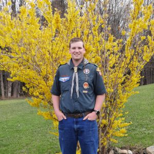 a young man wearing a Scout uniform stands in front of a yellow shrub