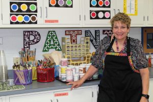 a woman with short hair, wearing a black apron stands in front of art supplies