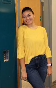a young woman with dark hair wearing a yellow shirt leans against a blue locker in a school hallway