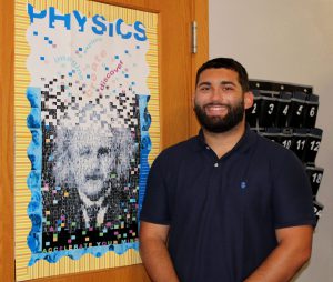 a man with facial hair and a short sleeve blue shirt stands in front of a physics poster with a picture of Albert Einstein
