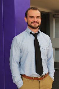 a man with facial hair, wearing a button down shirt and a dark tie stands in front of a purple wall