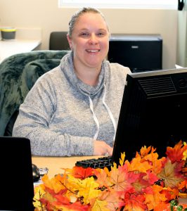 a blonde haired woman wearing a grey shirt sits behind a computer screen with fall leaves decorating the desk