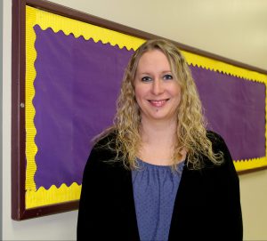 a woman with blond hair, wearing a blue shirt and black sweater, stands in front of a purple and gold bulletin board