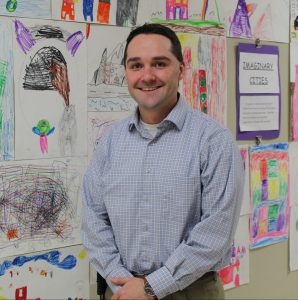 a man wearing a blue button down shirt stands in front of a bulletin board full of children's drawings