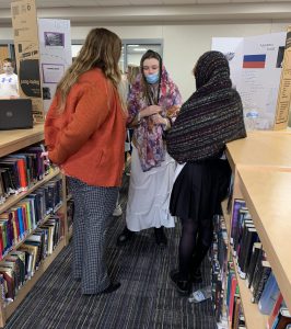an adult woman talks with two students about their class project in the library