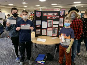 a group of four boys wearing masks stand in front of their group project in the library