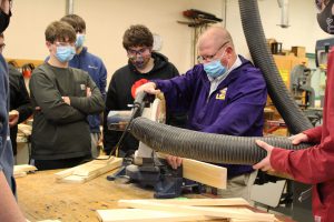 a man wearing a purple jacket, face mask and safety goggles demonstrates how to use a saw to his students