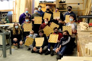 a group of high school aged students wearing face masks pose with their teacher, each holding up a checker board that they completed as a technology project