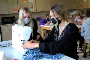 two female students wearing face masks make a bag using a sewing machine