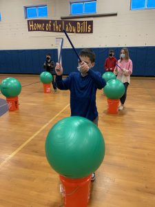 a boy in a gymnasium hits an exercise ball with drumsticks