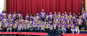 approximately 50 young students pose with three adults on a stage with a red curtain behind them. They are all wearing purple team t-shirts.