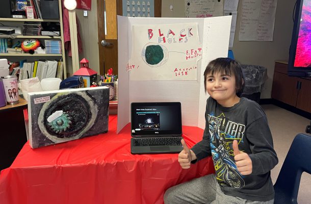 a young boy gives two thumbs up as he stands in front of his science project