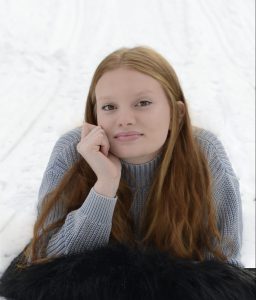 a young woman with long red hair and a grey sweater poses in front of a white backdrop