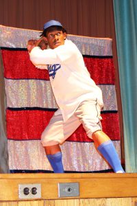 an actor portrays Jackie Robinson in a play and is shown swinging an imaginary bat, wearing a baseball uniform