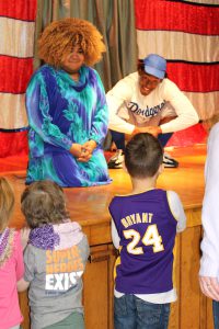three elementary students ask questions of two actors up on a theatrical stage 