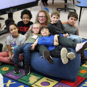 a woman wearing glasses sits in an inflatable chair with elementary aged students posing around her