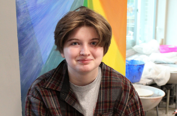 a student wearing a plaid flannel shirt sits in front of a rainbow colored wall