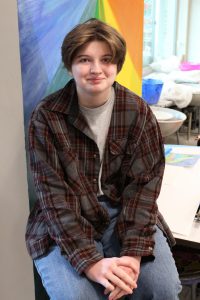 a teenager wears a plaid flannel shirt and sits in front of a colorful wall
