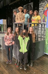 teenaged members of a theatrical crew pose on a piece of the set to a play, on a stage