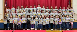 a group of about 50 elementary students in grey t-shirts pose in front of a red curtain