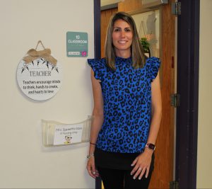 a woman wearing a blue and black shirt with black pants stands in the doorway of a classroom