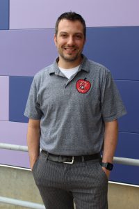 a man with dark hair and facial hair wearing a grey shirt and pants stands outside in front of a purple wall