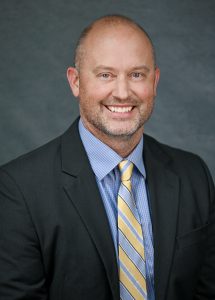 a man wears a dark suit, with a blue shirt and a yellow tie