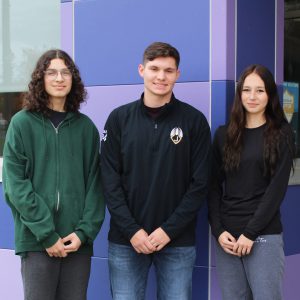two male students and one female student pose outside their school, in front of a purple wall