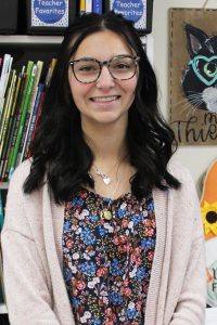 a woman with dark hair and glasses poses in front of a book shelf in a school