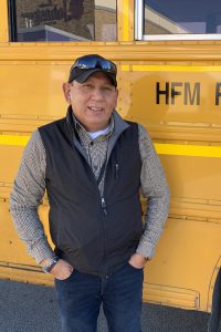 a man wearing a black vest poses next to a yellow school bus