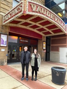 two teenagers, a boy and a girl, pose outside of a theater sign