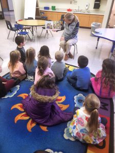 a woman reads a story to a classroom full of young children sitting on a blue carpet in a classroom
