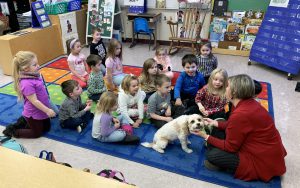 a woman with a little dog sit on the ground on a carpet in a classroom full of young children