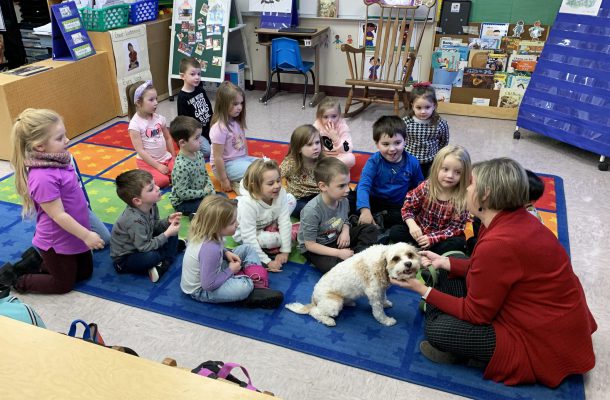a woman with a little dog sit on the ground on a carpet in a classroom full of young children