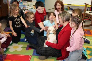 a dog sits in a woman's lap as young children sitting on the ground pet the dog