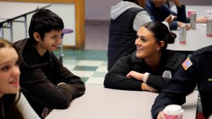 a woman kneels down to speak with a student who is sitting at a table in a cafeteria