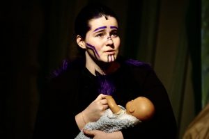 a young woman with face paint holds a fake baby on a theatrical stage