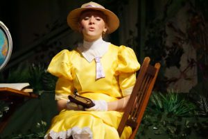 a young woman wears a yellow dress and hat on a theatrical stage
