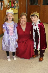 a woman wearing a maroon dress and wearing a tiara poses with two young students dressed as a king and a princess