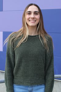 a woman with long hair wearing a green sweater stands outside in front of a purple wall