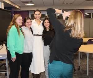 a woman takes a picture of another woman wearing a white dress, posing with three high school aged girls