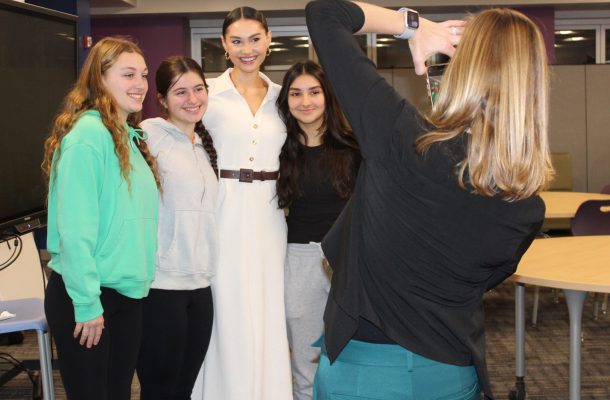 a woman takes a picture of another woman wearing a white dress, posing with three high school aged girls